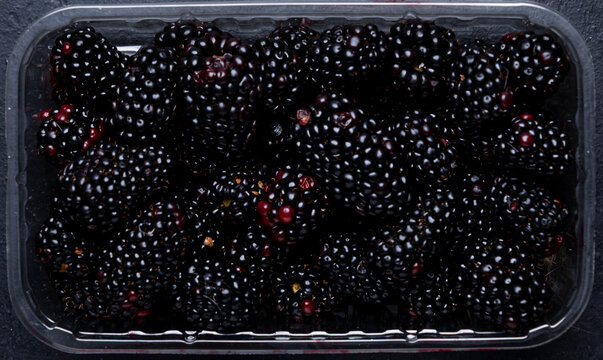 Fresh ripe blackberries in plastic punnet resting on dark surface. Supermarket berry packaging retail concept.