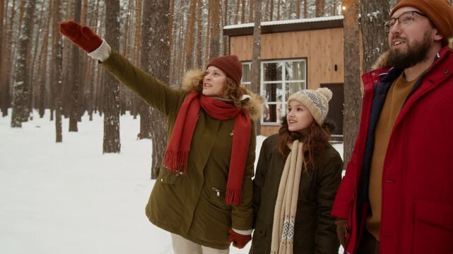 Caucasian family with middle aged parents and child girl standing in snowy forest near cabin, pointing and smiling. Ideal for winter holiday campaigns and travel agency promotions