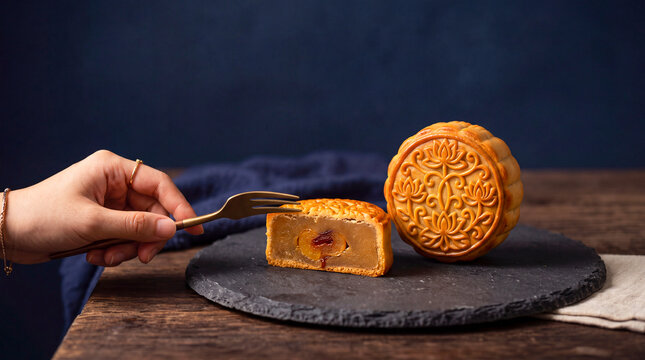 Mooncake Delight: A close-up shot captures a hand holding a fork, ready to sample a delectable mooncake, with another whole mooncake displayed beside it on a dark slate.