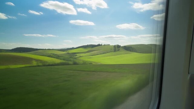 Sunlit View of Rolling Green Hills and Fields Passing by a Fast Moving Train Window under Blue Sky
