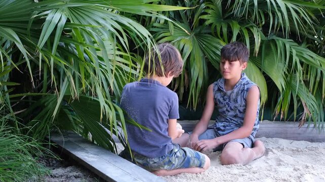 Two boys sit together on white sand beneath lush tropical palm foliage in a peaceful garden setting. The candid moment conveys friendship, communication, childhood, and emotional connection in nature.