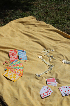 Various board games&mdash;such as a lotto set with cards and barrels, dominoes, and playing cards&mdash;are laid out on a yellow picnic blanket.