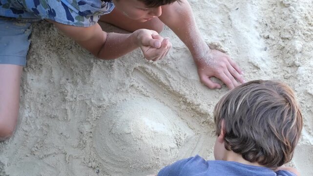 Two children work together to shape a sand volcano on white sand in an outdoor play area.