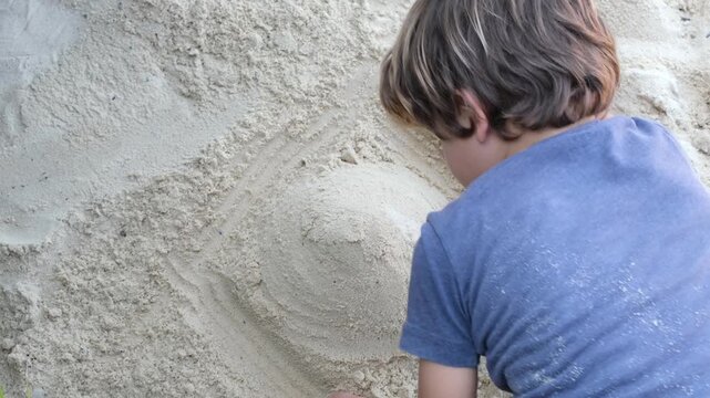 A young boy shapes and smooths a sand sculpture while playing outdoors in a tropical setting. The close-up captures creativity, imagination, and hands-on childhood exploration through natural play.
