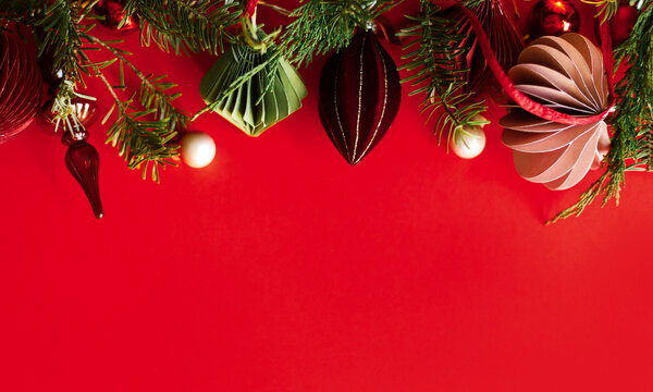 Christmas background with paper ornaments, glass finial, red baubles, and evergreen top border on red paper. Xmas card with copy space. Happy holidays
