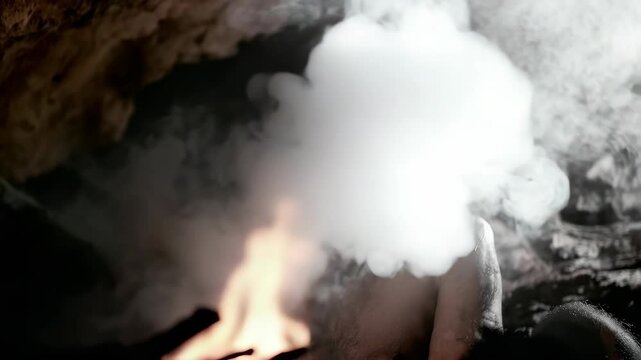 A man sits in a dark cave with smoke and steam coming from his mouth. He is bare-chested and appears to be in deep thought or distress. The cave environment suggests an eerie, moody atmosphere.