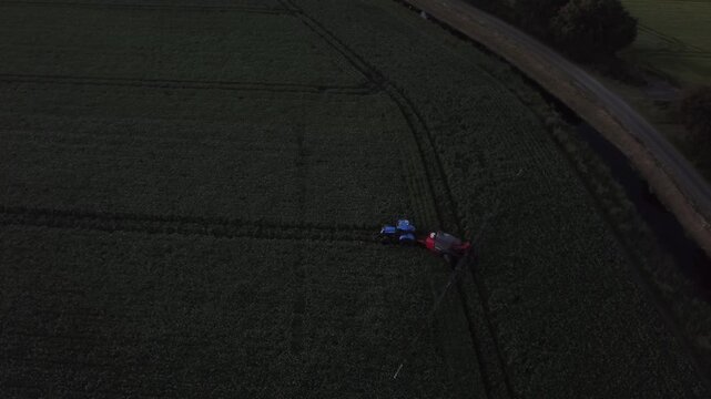 Aerial drone video capturing a blue tractor moving through a field, actively spraying pesticide or fertilizer onto rows of young green crops during the evening. As dusk settles, the sky darkens