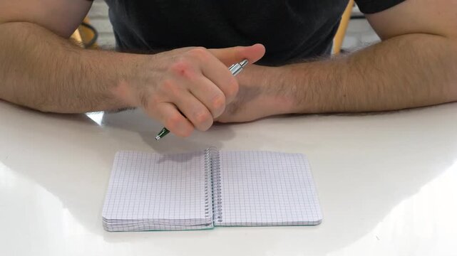 A man sitting at a table, holding a pen, with an open notebook whose pages are blank. He is twirling the pen in his hands as if to write.