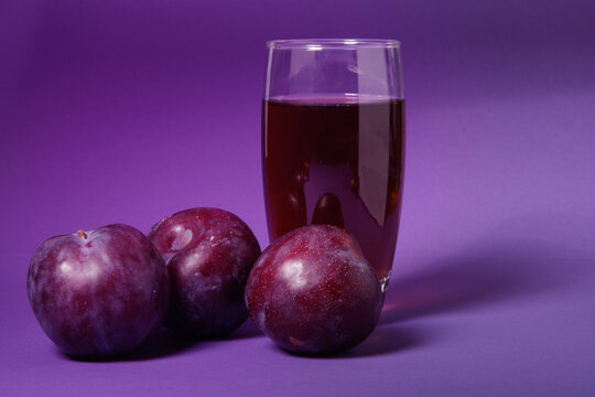Vaso de zumo de ciruela natural acompa&ntilde;ado de ciruelas frescas sobre un fondo morado. Concepto de alimentaci&oacute;n saludable, bebida natural, fruta fresca y fotograf&iacute;a de producto en estudio.
