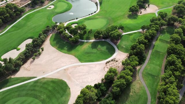 Aerial drone view of a lush green golf course landscape featuring pristine fairways white sand bunkers challenging water hazards winding paths and beautiful mature trees during bright sunny summer day