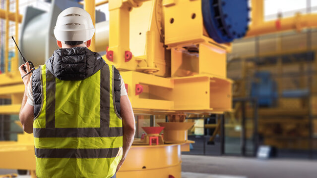 Industrial equipment. Worker uses radio communication while supervising heavy machinery operation, maintenance and safety at a manufacturing facility.