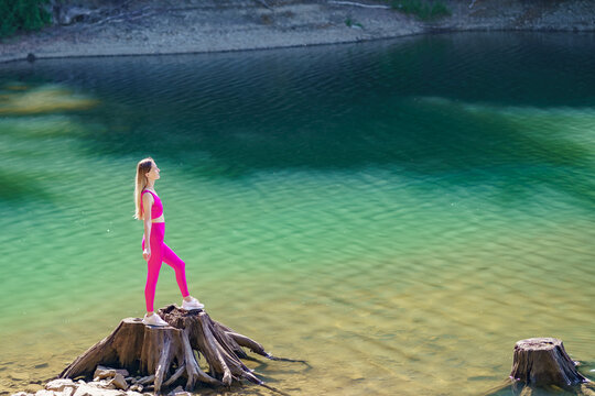Fit woman standing confidently on a tree stump above clear lake water in a scenic forest landscape. Concept of personal growth, confidence, achievement and healthy living