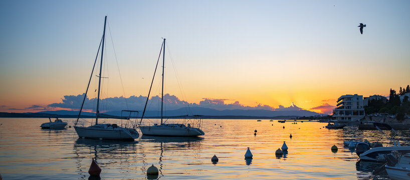 Sailboats floating on calm waters during sunset in Crikvenica, Croatia. Golden sky, colorful reflections and a flying seagull create a classic Mediterranean seascape