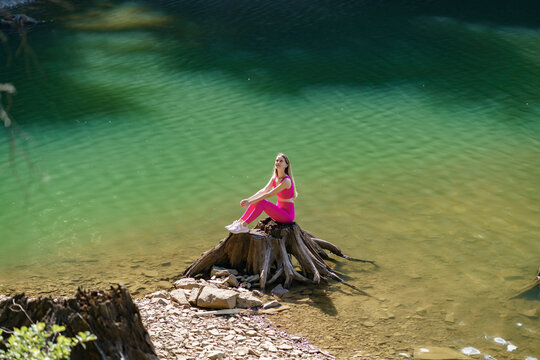 Young woman practicing meditation on a tree stump surrounded by clear green water in a tranquil natural environment. Concept of mental health, stress reduction and holistic wellbeing
