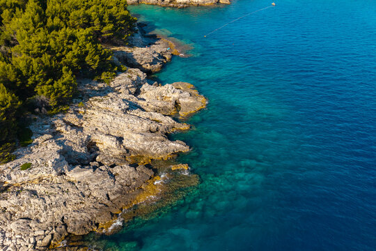 Top-down aerial view of crystal-clear turquoise water, natural sea caves, limestone cliffs, and boats anchored in the protected cove near Pula, Croatia