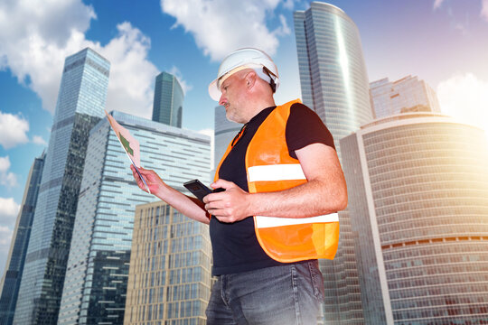 Construction inspection. Engineer reviews building plans and uses a smartphone while supervising modern high-rise construction, engineering and project development.