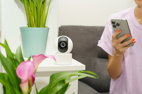 Indoor smart security camera placed in a modern home while a woman uses a smartphone nearby. Houseplants and a comfortable living area create a bright domestic setting