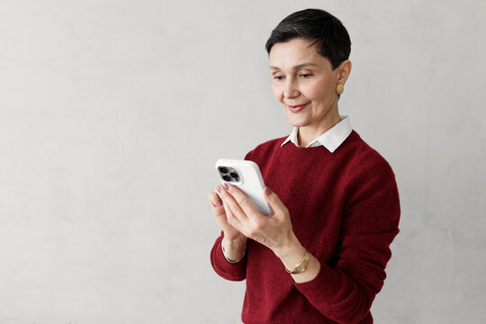 Middle-aged stylish woman entrepreneur in red jumper chatting with ai via smartphone application, using prompts to write content for social media promotion isolated on gray copy space background