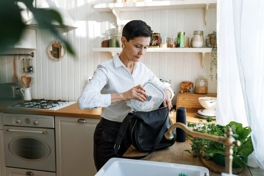 Side view of attractive mature Caucasian woman office worker in formal wear standing at kitchen in front of window and putting lunch box in her bag, before going to office. Healthy diet