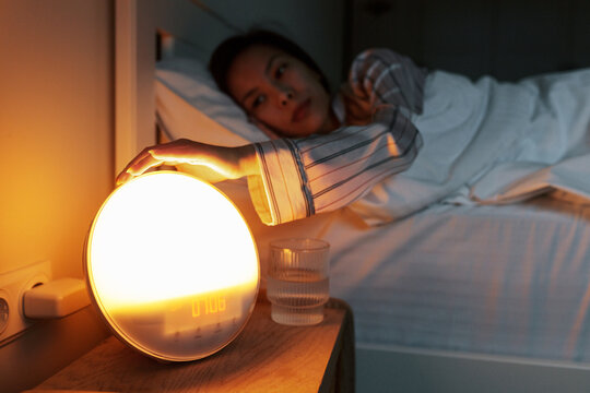 Hand of asian woman pushing stop button on round-shaped light alarm clock with dawn simulation with function of gentle waking up while laying in bed in early morning after having healthy sleep
