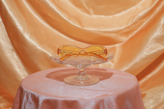 A pair of translucent jelly frame sunglasses sits on a clear glass stand against an orange satin background, highlighting the vibrant colors and unique materials of the eyewear