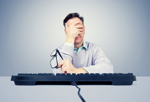 Frustrated man sitting at a computer keyboard, making a facepalm gesture with one hand while holding eyeglasses in the other. Concept of error, failure, frustration and work problems or online.