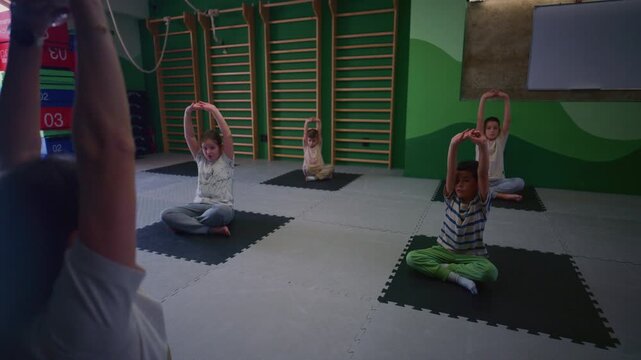 A female instructor leads a group of children through stretching and yoga exercises. Young kids participate in a supervised fitness class, focusing on flexibility and movement.