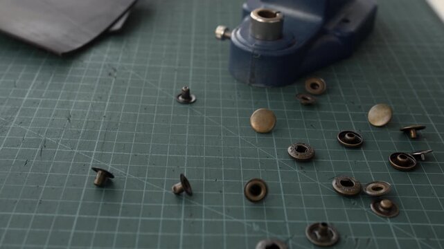Leather rivets and snap fasteners falling onto a green cutting mat in slow motion with a grommet machine in the background