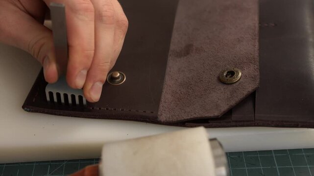 Artisan leather worker punching stitch holes in a brown handmade wallet using a stitching chisel and mallet in a workshop