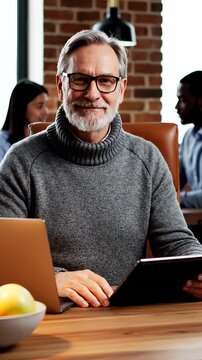 Confident senior man works with tablet and laptop at wooden desk. Mature businessman wearing glasses and sweater sits in coworking office. Colleagues work near brick wall in background.