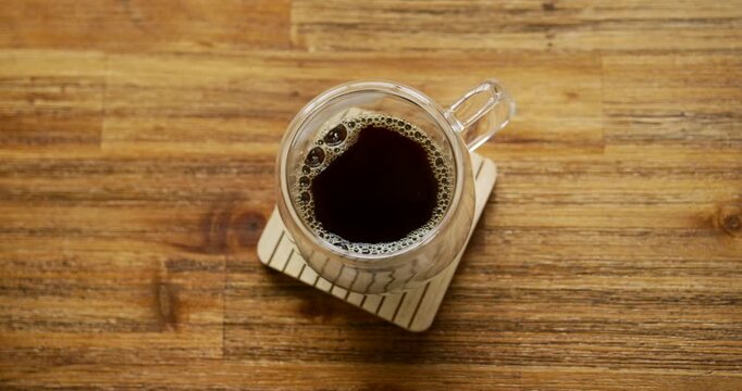 Placed clear glass mug of black coffee is settling on wooden coaster, bubbles shifting clockwise
