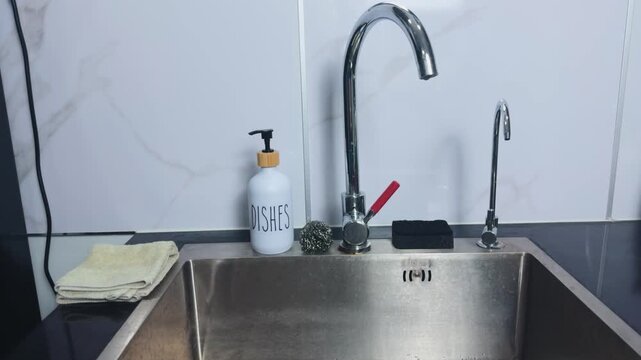 Modern kitchen sink featuring stainless steel basin, tall chrome faucet, dish soap dispenser, scrubbers, and neatly folded towel on dark countertop, ready for daily chores and hygiene