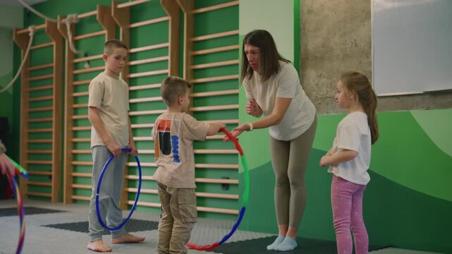 Children engage in a fun hula hoop exercise session with a female instructor in a bright gym. The young students participate in physical activity, showcasing development and teamwork.