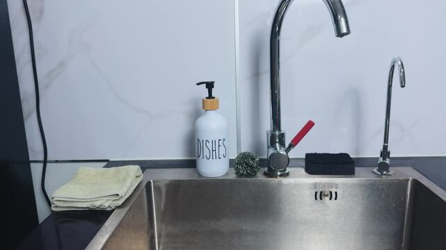 Kitchen sink area showing a soap dispenser for dishes, a scrubbing sponge, metal scourer, and a chrome faucet against a white tiled wall, representing household chores and daily cleaning