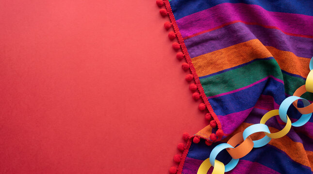 Festive background for Cinco de Mayo featuring a colorful striped serape blanket with pom-poms and a paper chain garland on a bright red background with copy space.