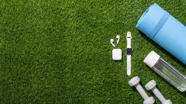 Top view flat lay of fitness equipment including a blue yoga mat, dumbbells, water bottle, smartwatch, and wireless earbuds arranged on green grass turf background.