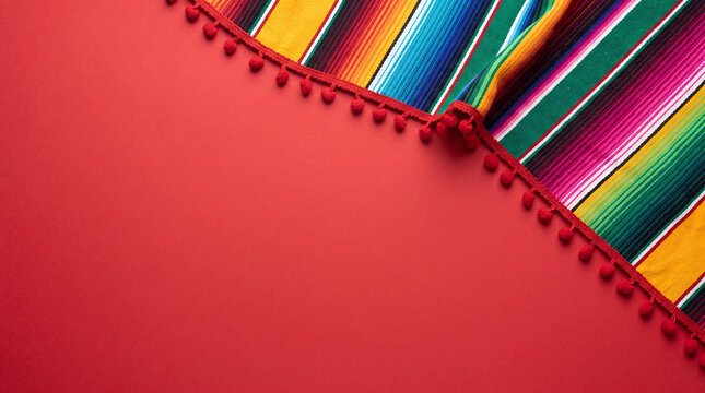 Festive background featuring a colorful striped Mexican serape blanket and red pom pom trim on a vibrant red surface with copy space.