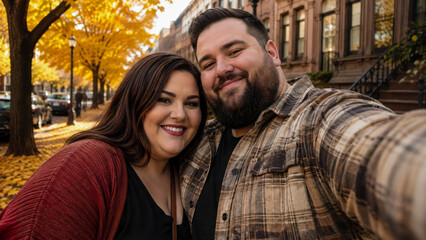 Fototapeta premium Happy plus-size couple taking selfie together on beautiful tree-lined street in city during vibrant autumn day with golden fall foliage and city buildings in background