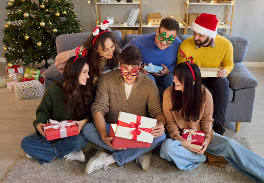 Christmas friends exchange gift with laughter and surprise. At home, friends unwrap gifts by the tree at a party, wearing Santa hats and festive glasses. Joyful holiday togetherness at home.