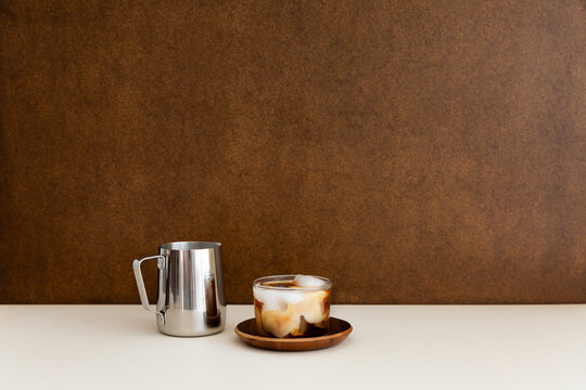 Un caf&eacute; cr&egrave;me glac&eacute; servi dans un verre transparent et pos&eacute; sur une soucoupe en bois sur une table blanche, un pot &agrave; lait en inox pos&eacute; &agrave; c&ocirc;t&eacute;, d&eacute;grad&eacute; marbr&eacute; dans la tasse, fond brun en arri&egrave;re-plan