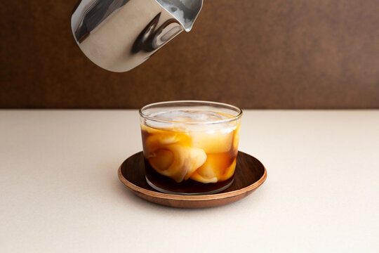 Caf&eacute; cr&egrave;me glac&eacute; en gros plan servi dans un verre transparent pos&eacute; sur une soucoupe en bois, pot &agrave; lait en inox en haut de l'image, fond brun en arri&egrave;re-plan, m&eacute;lange onctueux et d&eacute;grad&eacute; marbr&eacute;