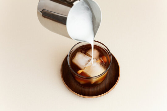 Un caf&eacute; cr&egrave;me glac&eacute; servi dans un verre transparent et pos&eacute; sur une soucoupe en bois, filet de lait qui coule d'un pot &agrave; lait en inox sur les gla&ccedil;ons et le caf&eacute;, vue du dessus en plong&eacute;e