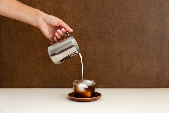 Caf&eacute; cr&egrave;me glac&eacute; servi dans un verre transparent pos&eacute; sur une soucoupe en bois, une main qui verse du lait avec un pot &agrave; lait en inox, filet de lait, d&eacute;grad&eacute; marbr&eacute;, arri&egrave;re-plan brun
