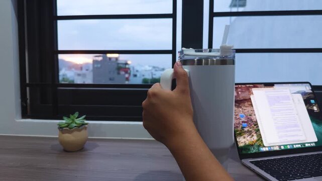 Person's hand holding a reusable insulated tumbler with a straw while working on a laptop at a desk with a succulent plant. Viewing the city skyline and sunset through a window