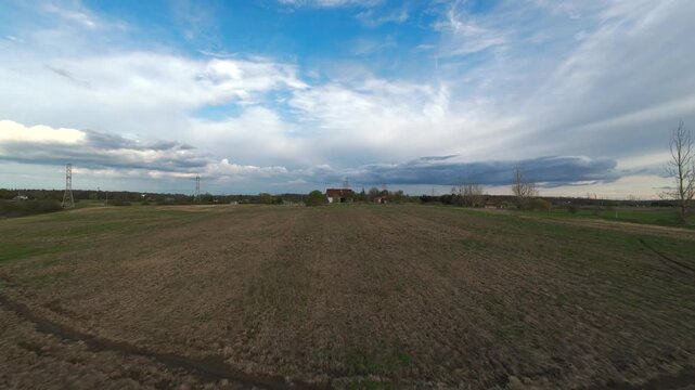 Aerial view moving forward across an expansive empty meadow of dry grass towards a distant red farmhouse and old rustic barns under a dramatic cloudy sky during spring season.