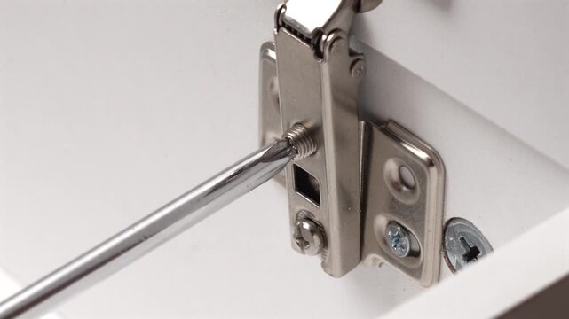 Close-up of Phillips screwdriver adjusting Euro-style cabinet hinge on white furniture during assembly or repair