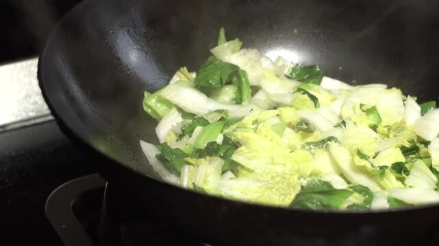 Stir-frying chopped napa cabbage and bok choy in a black carbon steel wok over gas flame