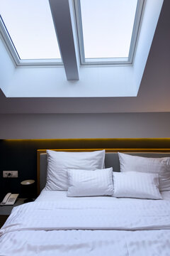 Close view of a neatly arranged hotel bed beneath large skylights, featuring crisp white pillows, striped bedding, a dark accent wall and warm indirect lighting