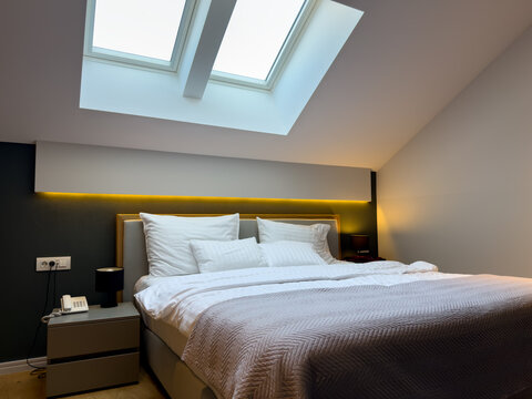 Bright contemporary hotel bedroom with a large neatly made bed, white pillows and linens, warm ambient lighting, dark accent wall and skylights providing natural daylight