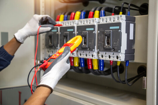 Industrial electrician holding digital clamp meter and using test probe to inspect commercial circuit breaker panel.